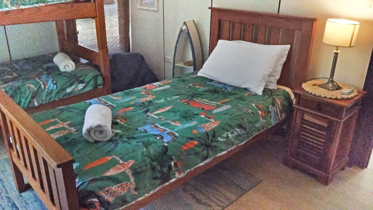 A single bed with a patterned bedspread, a wooden headboard, a bedside table with a lamp, and a rolled towel on the floor.