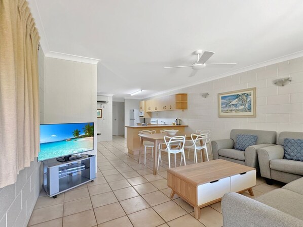 A modern living room with a flat-screen TV, a wooden coffee table, and a sofa. There is a kitchen area with a dining table and chairs.
