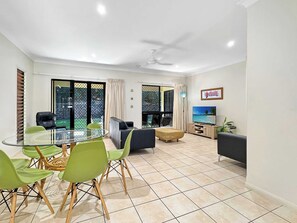 Uma sala de estar moderna com uma mesa de jantar de vidro, cadeiras verdes, um sofá, uma TV e uma varanda com vista.