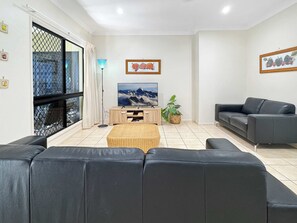 Una sala de estar moderna con un sofá de cuero negro, una mesa de centro de madera, un televisor sobre un mueble de madera y un ventanal con cortinas.