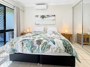 A hotel room with a bed, two bedside tables, a wall-mounted air conditioner, a large window with a view, and a tropical-themed artwork on the wall.