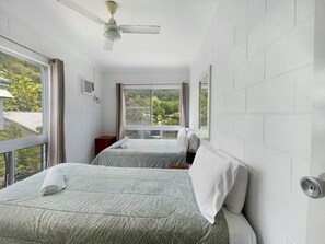 A room with two beds, a ceiling fan, a window with curtains, and a view of greenery outside. - Arcadia Beach Unit 4 (Arcadia)