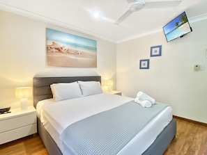 Ein ordentlich eingerichtetes Schlafzimmer mit einem Bett, Nachttischlampen, einem Deckenventilator, einem an der Wand montierten Fernseher und einem Gemälde mit Strandmotiv.