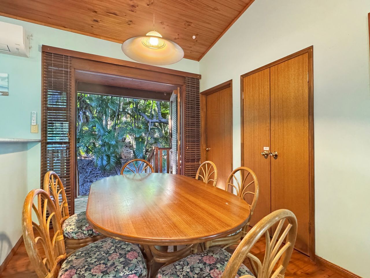 Uma sala de jantar com mesa e cadeiras de madeira, ventoinha no teto e vista para o verde lá fora.