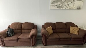 Deux canapés marron avec des coussins, un sac à dos posé sur le canapé de gauche et une œuvre d’art encadrée accrochée au mur.