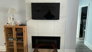 Un salon avec un téléviseur à écran plat fixé au mur, qui est recouvert de marbre, un meuble en bois doté de portes en verre et une cheminée située en dessous.