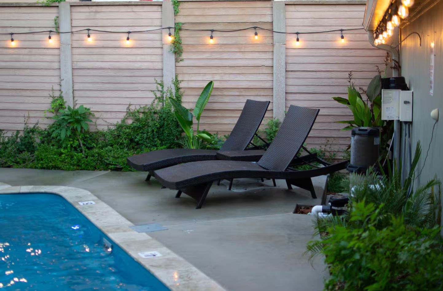 A poolside area with lounge chairs, a wooden fence, string lights, and greenery.