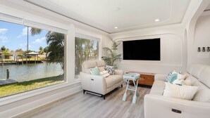 Amplia sala de estar con un ventanal con vistas a un cuerpo de agua, un sofá blanco, una pequeña mesita auxiliar y un televisor de pantalla plana montado en la pared.