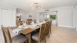 Un comedor con una larga mesa de madera dispuesta para seis comensales, rodeada de sillas de madera con asientos acolchados. Un moderno candelabro cuelga del techo, y la habitación tiene paredes blancas y un piso de baldosas.