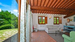 A room with a brick floor, wooden ceiling beams, a white sofa, a green armchair, and a window overlooking a garden. - Francigena 6, Castelfiorentino, Italy (Castelfiorentino)