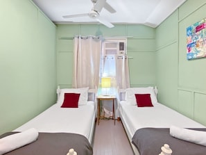 Une chambre double avec deux lits, un ventilateur de plafond, une fenêtre avec des rideaux et une petite table.