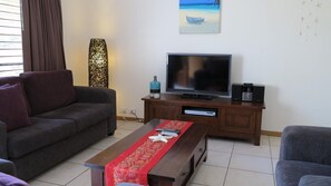 A living room with a sofa, a coffee table with a red cloth, a TV on a wooden stand, and a decorative wall piece.