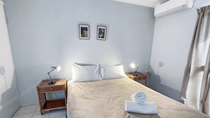 A hotel room with a bed, two bedside tables with lamps, a wall-mounted air conditioner, and a window with curtains. - Magnetic Shores Unit 2 (Arcadia)