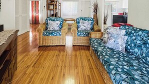 A spacious living room with a wooden floor, two wicker sofas with floral upholstery, a ceiling fan, and a view into the adjacent dining area.