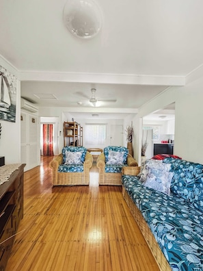 A spacious living room with a wooden floor, two wicker sofas with floral upholstery, a ceiling fan, and a view into the adjacent dining area.