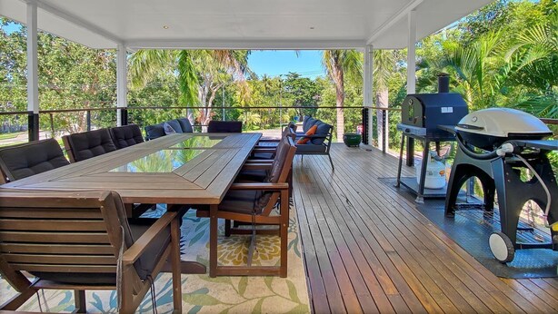 An outdoor covered patio with a long wooden table and chairs, a grill, and a view of lush greenery.