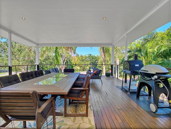 Une terrasse extérieure couverte, avec une longue table et des chaises en bois, un barbecue et une vue sur une végétation luxuriante.