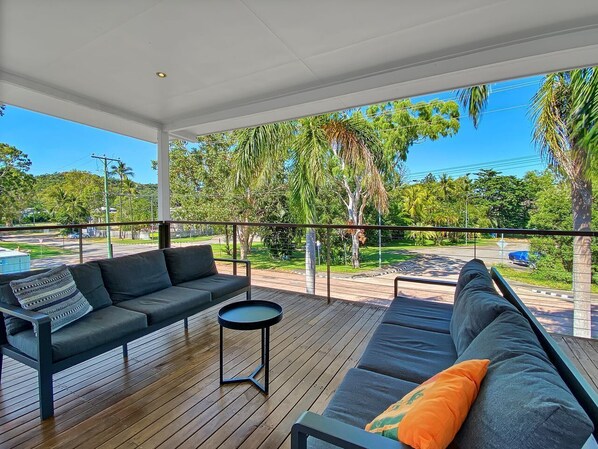 A covered outdoor seating area with a sofa and a small table, overlooking a street and greenery.