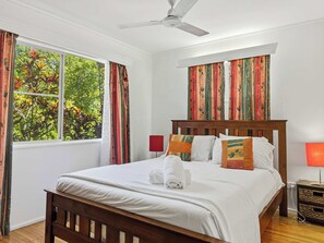 Uma cama bem arrumada com roupa de cama branca e cabeceira de madeira, um ventilador de teto e uma janela com cortinas.