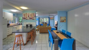 Eine moderne Küche mit Kochinsel, Barhocker, einem Esstisch mit blauen Stühlen und einer Sitzecke mit Sofa und Kissen.
