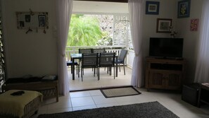 A living room with a TV, a sofa, a dining table with chairs, and a balcony with a view of greenery.