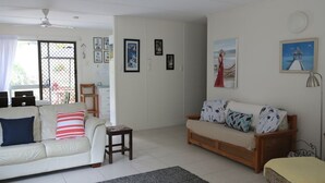 Ein Wohnzimmer mit einer Couch, einem kleinen Tisch und einem Korbsessel. An den Wänden hängen gerahmte Bilder.