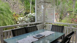 Ein Tisch mit Glasplatte und Unterlage, umgeben von Stühlen, befindet sich auf einem Balkon mit weißer Brüstung und üppiger Bepflanzung im Hintergrund.