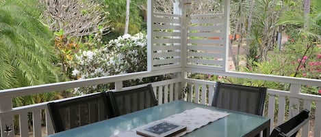 A glass-top table with a placemat, surrounded by chairs, set on a balcony with a white railing and lush greenery in the background.