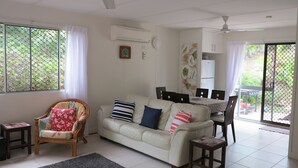 Ein Wohnzimmer mit einer Couch, Sesseln, einem Esstisch mit Stühlen, einer Küchenzeile und einer Glastür zum Außenbereich.