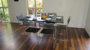 A dining area with a table set for four, surrounded by metal chairs, large windows with a view of greenery, and a wooden floor.