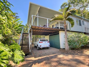 A white SUV parked under a covered garage, a staircase leading to a second level, and a palm tree in the foreground.