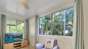 A modern living room with a flat-screen TV, a sofa with a beach-themed pillow, a blue ottoman, and large windows with a view of palm trees.
