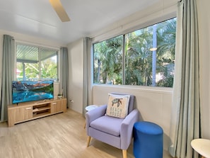A modern living room with a flat-screen TV, a sofa with a beach-themed pillow, a blue ottoman, and large windows with a view of palm trees.
