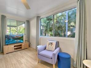 A modern living room with a flat-screen TV, a sofa with a beach-themed pillow, a blue ottoman, and large windows with a view of palm trees.