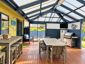 Une terrasse extérieure avec une table et des chaises en bois, un barbecue et un espace abrité doté d’un toit en métal.