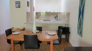 Een eetgedeelte met een houten tafel en stoelen, een keuken met witte kasten, en een schilderij aan de muur.