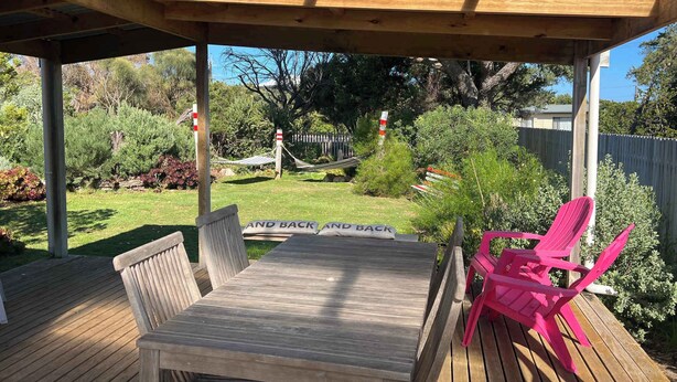 A wooden deck with a table and chairs, surrounded by a fenced yard with trees and a hammock.
