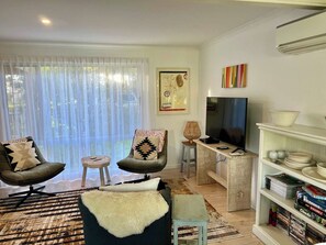 Un salon comprenant deux fauteuils, une table basse, un téléviseur, une bibliothèque et une grande fenêtre ornée de rideaux légers.