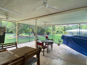 Een overdekt terras met een tafel, stoelen, een plafondventilator en uitzicht op een achtertuin.