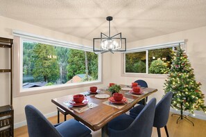 A dining area with a wooden table set for four, large windows offering a view of trees and a lake, and a decorated Christmas tree. - Relaxing Lakeside Bungalow,Steps to Lake Samish Park to enjoy lake activities (Bellingham)