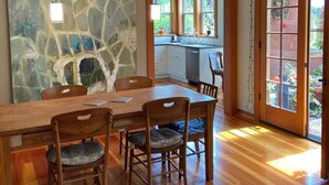Un comedor con una mesa grande y sillas, una pared de piedra con arte abstracto, y vista a una cocina a través de una puerta de vidrio.