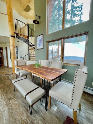 A spacious dining area with a wooden table and upholstered chairs, a staircase, and a large window with a view of trees.