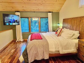 A bedroom with a wooden ceiling, a large bed with white bedding, a TV mounted on the wall, and a window with curtains. - Mountain Getaway- Hot Tub, Hiking, 5mins to Ski Resort (Durango)