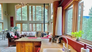 Un acogedor interior de cabaña con una mesa de comedor de madera, un ventanal con cortinas, un lavamanos empotrado y una zona de estar con sofá y sillas.