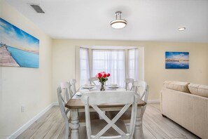 A dining area with a table set for four, white chairs, a vase with flowers, and a painting of a beach scene on the wall.