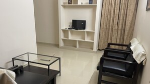 A living room with a glass-top coffee table, a black sofa, a black armchair, and a white shelving unit against the wall.