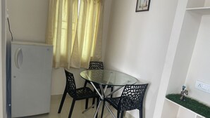 A small dining area with a glass table and four chairs, a white refrigerator, and a window with yellow curtains.