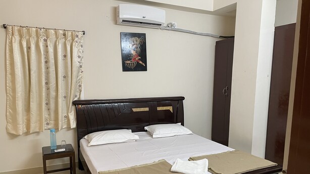 A neatly made bed with white linens, a small bedside table with a bottle and glass, a wall-mounted air conditioner, a framed artwork, and a wardrobe.