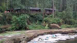 Een chalet met een terras dat uitkijkt over een bebost gebied, een rotsachtige kustlijn met een klein steiger en een paar stoelen en een kajak.