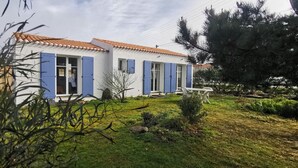 Edifici blanc amb persianes blaves, teulada de rajola i un petit jardí amb gespa i arbustos.